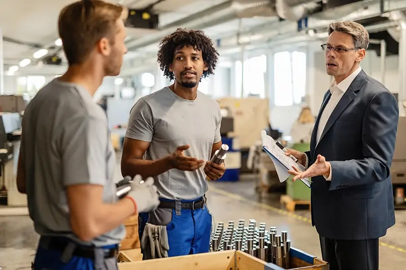 Operator met gele helm bespreekt productie met manager op de fabrieksvloer; lean cultuur op de werkvloer en Leader Standard Work
